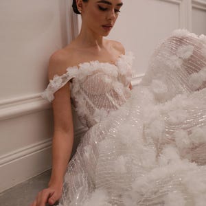 Op de afbeelding: Een witte off-the-shoulder trouwjurk met bloemmotieven en een zwierige rok. De jurk heeft een doorschijnend lijfje met ingewikkelde details en is versierd met sprankelende accenten. De bruid zit tegen een witte muur.
