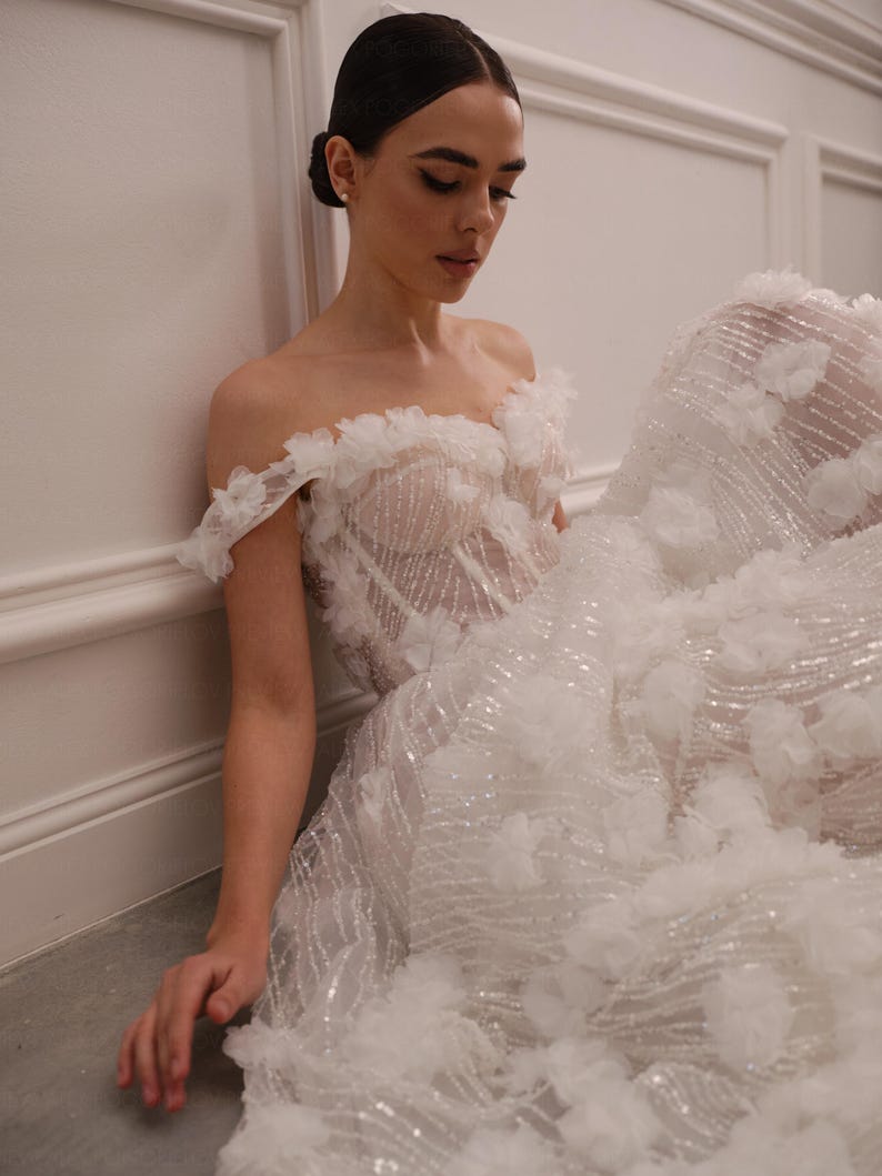Op de afbeelding: Een witte off-the-shoulder trouwjurk met bloemmotieven en een zwierige rok. De jurk heeft een doorschijnend lijfje met ingewikkelde details en is versierd met sprankelende accenten. De bruid zit tegen een witte muur.