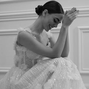 Op de afbeelding: Een zwart-wit afbeelding van een formele jurk met bloemversieringen en sprankelende details. De jurk heeft een nauwsluitend lijfje en een zwierige rok. Het model lacht met gevouwen handen.