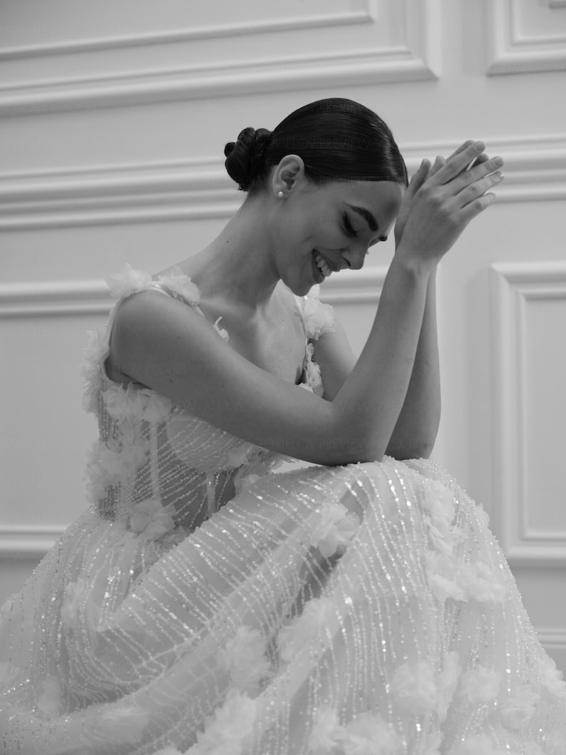 Op de afbeelding: Een zwart-wit afbeelding van een formele jurk met bloemversieringen en sprankelende details. De jurk heeft een nauwsluitend lijfje en een zwierige rok. Het model lacht met gevouwen handen.