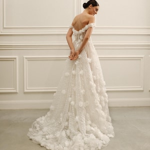 Op de afbeelding: Een witte off-the-shoulder trouwjurk met bloemendetails. De jurk heeft een zwierige rok en een gedetailleerd lijfje. De jurk is versierd met driedimensionale bloemenapplicaties, wat een textuur en romantische uitstraling cre&euml;ert. De jurk is vloerlang en heeft een sleep.