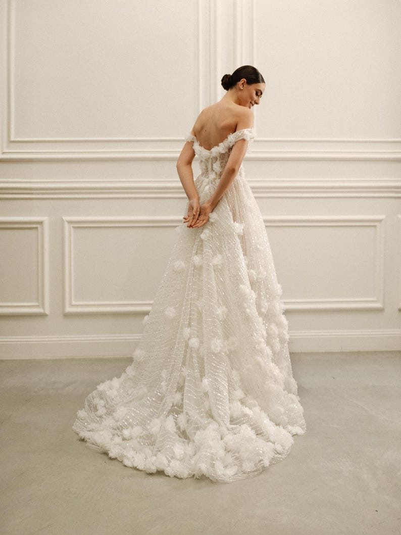 Op de afbeelding: Een witte off-the-shoulder trouwjurk met bloemendetails. De jurk heeft een zwierige rok en een gedetailleerd lijfje. De jurk is versierd met driedimensionale bloemenapplicaties, wat een textuur en romantische uitstraling cre&euml;ert. De jurk is vloerlang en heeft een sleep.