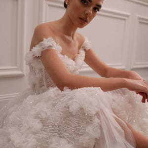 Op de afbeelding: Een witte off-the-shoulder trouwjurk met bloemversieringen en een zwierige rok. De jurk heeft ingewikkeld kralenwerk en een transparante overlay, wat een romantische en elegante uitstraling cre&euml;ert. De jurk is ontworpen voor een speciale gelegenheid.