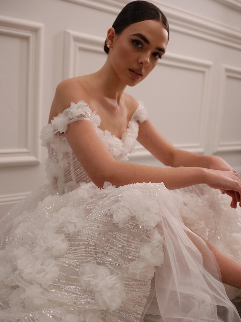 Op de afbeelding: Een witte off-the-shoulder trouwjurk met bloemversieringen en een zwierige rok. De jurk heeft ingewikkeld kralenwerk en een transparante overlay, wat een romantische en elegante uitstraling cre&euml;ert. De jurk is ontworpen voor een speciale gelegenheid.