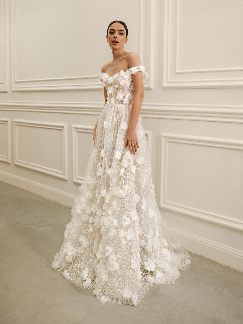 Op de afbeelding: Een witte off-the-shoulder trouwjurk met bloemenversieringen. De jurk heeft een nauwsluitend lijfje en een zwierige rok, versierd met 3D bloemenapplicaties. De jurk is gemaakt van een glinsterende stof, wat een romantische en elegante uitstraling cre&euml;ert.