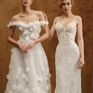 Op de afbeelding: Twee witte trouwjurken. E&eacute;n jurk heeft een off-the-shoulder halslijn met bloemversieringen en een volle rok. De andere jurk heeft een nauwsluitend lijfje met spaghettibandjes en een kolomrok, eveneens versierd met bloemendetails.