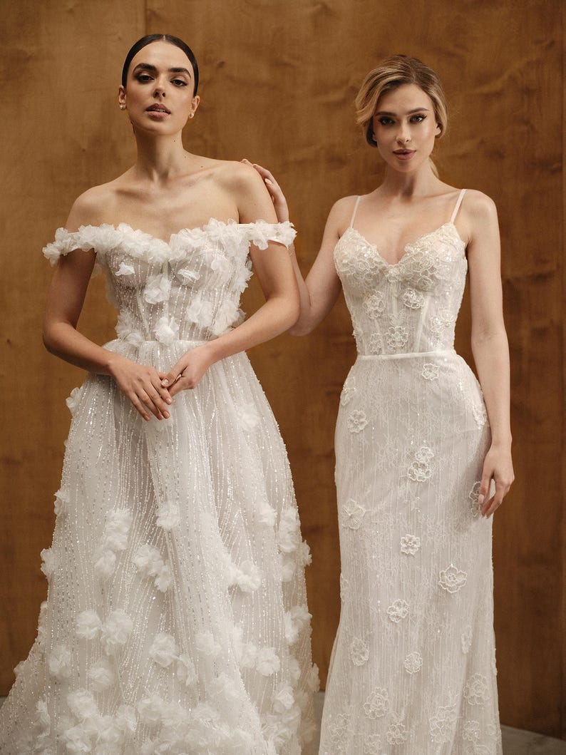 Op de afbeelding: Twee witte trouwjurken. E&eacute;n jurk heeft een off-the-shoulder halslijn met bloemversieringen en een volle rok. De andere jurk heeft een nauwsluitend lijfje met spaghettibandjes en een kolomrok, eveneens versierd met bloemendetails.