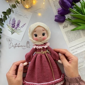 May include: A handmade crochet doll with a burgundy dress, white lace trim, and a cream-colored head covering. The doll has rosy cheeks and is held by hands. The background includes purple tulips and a newspaper.