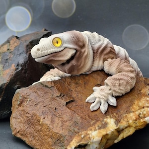 May include: A detailed sculpture of a lizard with a smiling expression, in cream and brown tones. The lizard rests on a textured brown rock, with bright yellow eyes. The background is dark with blurred light orbs.
