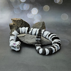 May include: A detailed toy snake in shades of grey, white, and black, posed on a grey stone. The snake's head features purple eyes and an open mouth. The body is segmented with alternating white and black sections.