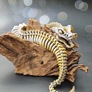 May include: A detailed, articulated snake sculpture with a skeletal design. The snake is primarily off-white with gold accents, posed on a piece of driftwood. The sculpture's open mouth reveals teeth, and the overall aesthetic is a blend of nature and fantasy.