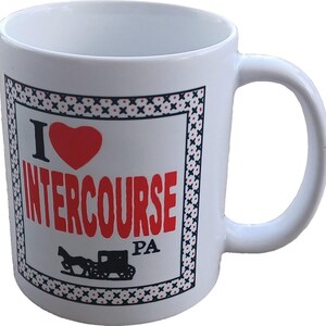Può includere: Tazza in ceramica bianca con manico. La tazza presenta la scritta "I ❤️ INTERCOURSE PA" in rosso, con un simbolo a cuore rosso e un'illustrazione di una carrozza trainata da cavalli nera. Il design è incorniciato da un bordo decorativo.