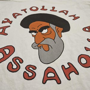 Op de afbeelding: Wit t-shirt met een cartoon illustratie van een man met een grijze baard en een zwarte hoed. De rode tekst "AYATOLLAH ASSAHOL" is in een cirkel rond de afbeelding gedrukt. Het cartoonpersonage heeft een oranje gezicht en een strenge uitdrukking.