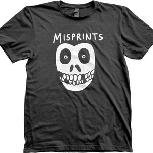 May include: Black t-shirt with a white graphic of a skull-like face and the word "MISPRINTS" above it. The face has large, round eyes and a toothy grin. The shirt is made of a soft material.