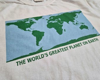 May include: Cream-colored t-shirt featuring a green world map design on a light blue background. The text "THE WORLD'S GREATEST PLANET ON EARTH" is printed in green below the map. The shirt is made of a soft, comfortable material.