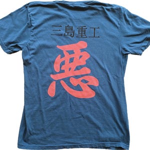 Puede incluir: Camiseta azul con caracteres japoneses estampados en la espalda. El texto superior dice "三島重工" en negro, y debajo hay un gran carácter rojo. La camiseta tiene mangas cortas y cuello redondo.