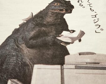 May include: White t-shirt featuring a Godzilla graphic holding papers near a printer. The word "Canon" is printed in red at the top left, with Japanese text on the right.