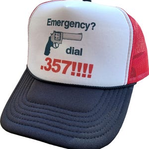 Puede incluir: Gorra de camionero con panel frontal blanco y laterales de malla roja. La parte delantera presenta el texto "Emergency?" sobre un dibujo de un revólver y las palabras "dial .357!!!!" en rojo.