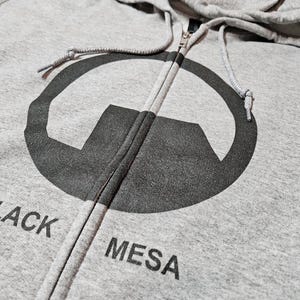 May include: A grey zip-up hoodie with a large black graphic and the words "BLACK MESA" printed beneath. The hoodie has a drawstring hood and a full-length zip. The design is centred on the chest.
