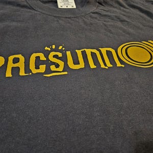 May include: Dark gray t-shirt with the word "PACSUN" in a distressed yellow font. The design includes a stylized sun graphic. The shirt is made of a soft material and has a crew neck.
