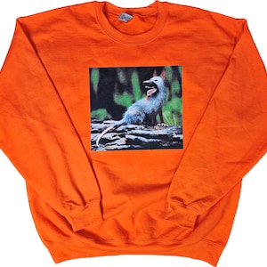 May include: Orange sweatshirt with a graphic of a blue opossum with its mouth open. The opossum is set against a green and black background. The sweatshirt has long sleeves and a crew neck.