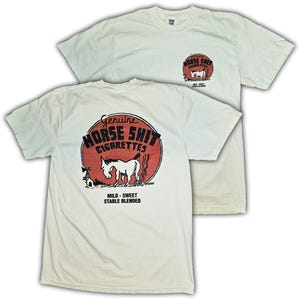 以下が含まれることがあります： オフホワイトの半袖Tシャツで、背面にグラフィック、左胸に小さめの同デザインがプリントされています。赤い円の中に馬のイラストと「Genuine HORSE SHIT CIGARETTES MILD - SWEET STABLE BLENDED」という文字が描かれています。