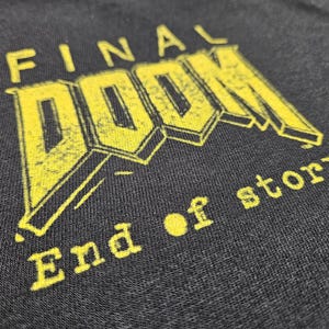 Puede incluir: Primer plano de una camiseta gris oscuro con las palabras "FINAL DOOM" en texto amarillo estilizado. Debajo del título, el texto "End of story." está impreso en amarillo. La camiseta tiene una apariencia texturizada.