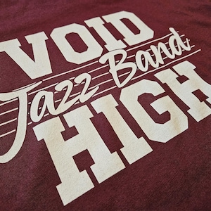 May include: A maroon t-shirt featuring white screen-printed text that reads VOID JAZZ BAND HIGH. The design includes a musical staff with a treble clef integrated into the lettering, creating a graphic layout across the front of the garment.