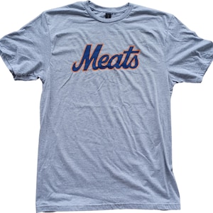 May include: A heather gray t-shirt with the word "Meats" in a blue script font with an orange outline. The shirt has short sleeves and a crew neck.