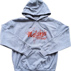 Puede incluir: Una sudadera con capucha gris con un bolsillo delantero y cordón. La sudadera presenta el texto rojo "Melsky's Auto Repair" con un contorno de mapa. La sudadera con capucha está hecha de un material suave y cómodo.