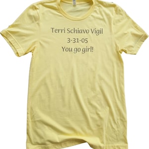 May include: A light yellow t-shirt with the text "Terri Schiavo Vigil 3-31-05 You go girl!" printed in a gray font. The shirt has short sleeves and a crew neck.