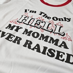 May include: White t-shirt with a red trim around the collar and sleeves. The shirt features the text "I'm The Only HELL My Momma Ever Raised" in black, with flames in red.
