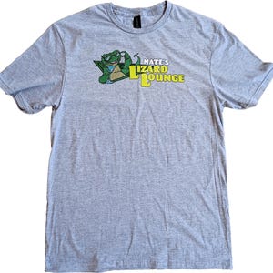 May include: A heather gray t-shirt featuring a cartoon lizard wearing sunglasses and holding a cocktail. The shirt has the text "Nate's Lizard Lounge" in yellow and green.