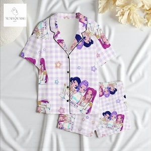 A pair of pajama bottoms with a cartoon-style design featuring multiple characters, including a girl with purple hair and a boy with a bow in his hair. The pajamas are displayed on a white background, and there are some flowers visible in the background.