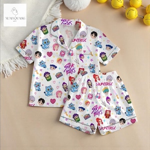 A children's pajama set with a colorful, cartoon-style print featuring various characters and designs. The pajamas are displayed on a beige background, along with a few yellow balls and a white blanket.