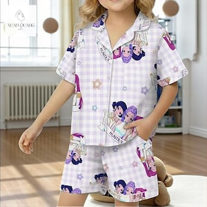 A young girl wearing a white and purple plaid pajama top with cartoon characters and a matching plaid skirt, standing in a room with a teddy bear.