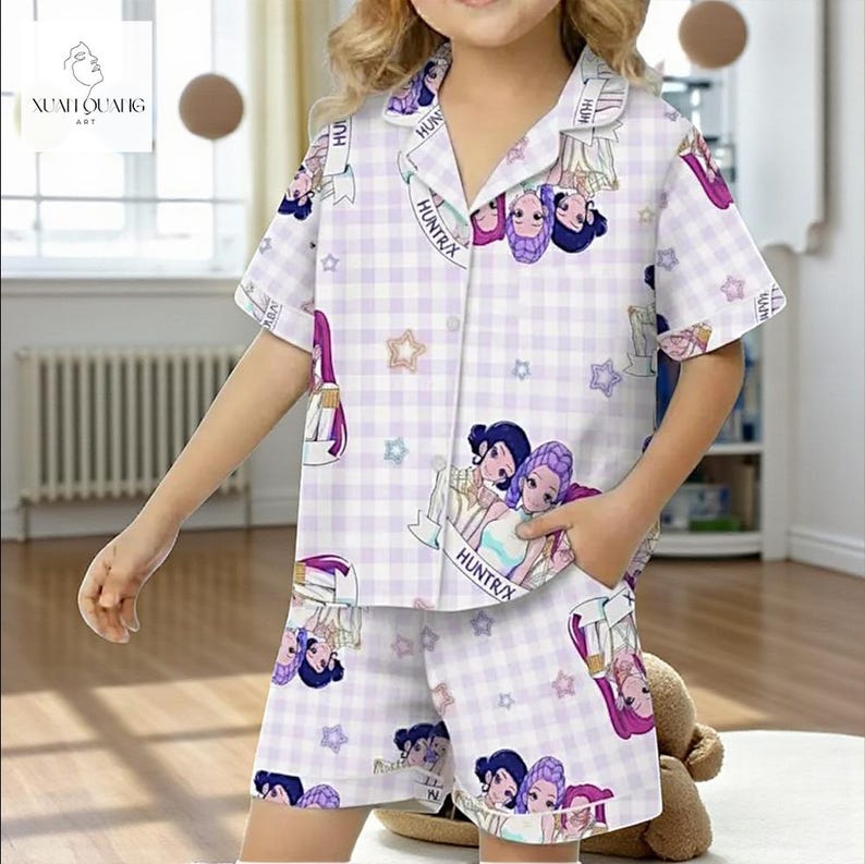 A young girl wearing a white and purple plaid pajama top with cartoon characters and a matching plaid skirt, standing in a room with a teddy bear.