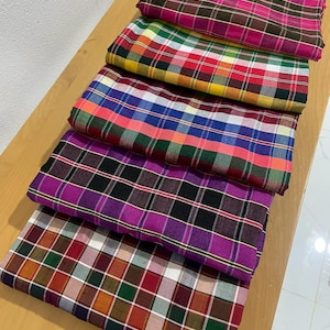 May include: A stack of six folded plaid fabric pieces in various colors. The fabrics feature a classic checkered pattern in shades of pink, brown, red, yellow, green, blue, purple, black, and white. The textiles are arranged on a light brown wooden surface.