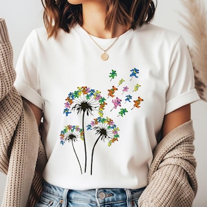 May include: White t-shirt featuring a dandelion design with colorful bear-shaped seeds blowing in the wind. The dandelion heads are multi-colored, and the bears are in various colors. The shirt is a casual, everyday wear item.