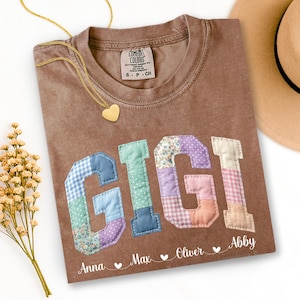 May include: Brown t-shirt with the name "GIGI" in patchwork letters. The shirt has the names "Anna", "Max", "Oliver", and "Abby" printed below. A gold heart necklace and a tan hat are also visible.