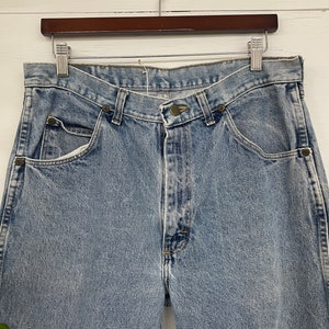 Op de afbeelding: Lichtblauwe spijkerbroek met een knoopsluiting en vijf zakken. De broek hangt aan een houten hanger.