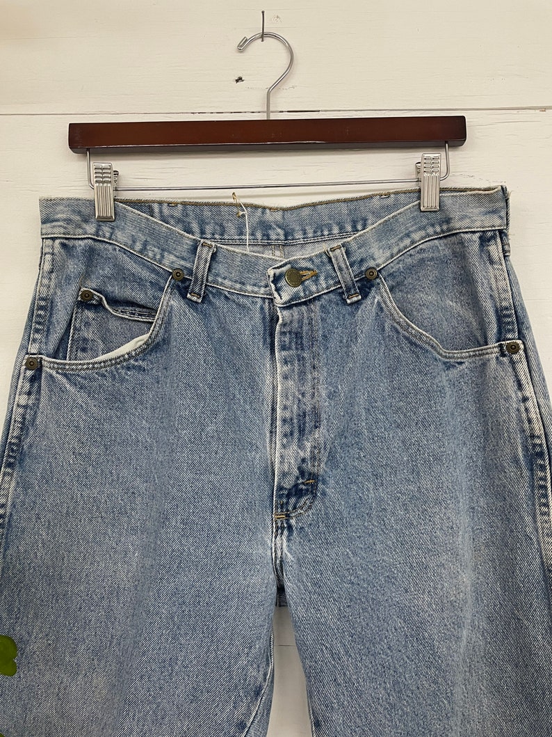 Op de afbeelding: Lichtblauwe spijkerbroek met een knoopsluiting en vijf zakken. De broek hangt aan een houten hanger.