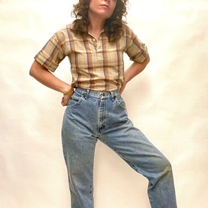 Op de afbeelding: Een vrouw draagt een geruit, kort mouwig shirt en een lichtblauwe, hoge taille jeans.