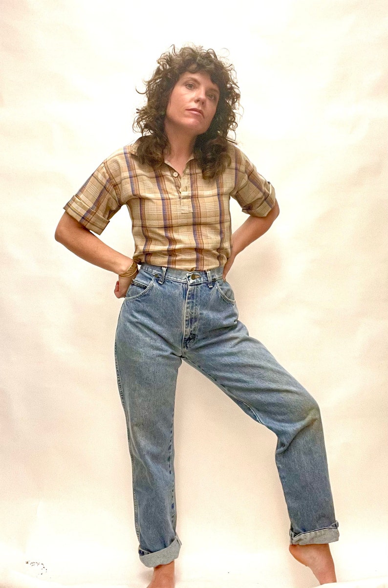 Op de afbeelding: Een vrouw draagt een geruit, kort mouwig shirt en een lichtblauwe, hoge taille jeans.