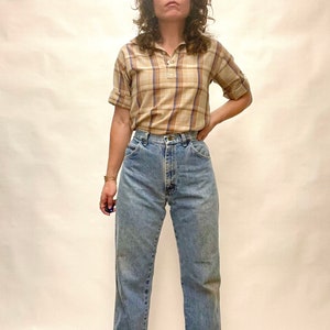 Op de afbeelding: Een vrouw draagt een lichtbruin en blauw geruit overhemd met korte mouwen, een hoge kraag en een ritssluiting. Ze draagt ook lichtblauwe jeans met een hoge taille.