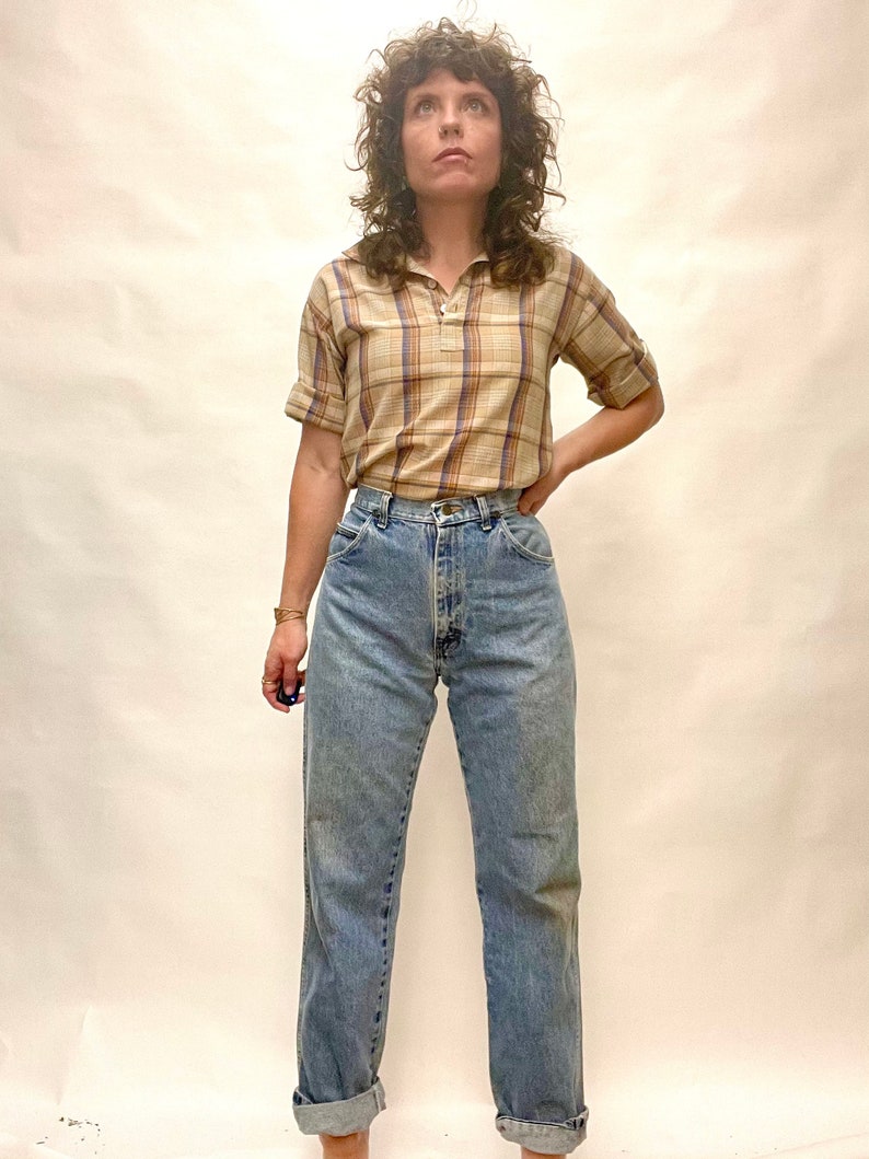 Op de afbeelding: Een vrouw draagt een lichtbruin en blauw geruit overhemd met korte mouwen, een hoge kraag en een ritssluiting. Ze draagt ook lichtblauwe jeans met een hoge taille.