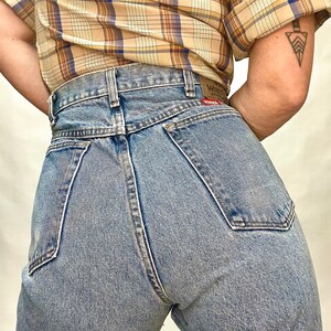 Op de afbeelding: Een lichtblauwe spijkerbroek met een hoge taille en een klassieke vijf-zak stijl. De spijkerbroek heeft een vervaagde wassing en een licht verweerde look. De achterzakken hebben een rood leren patch met het woord "Wrangler" erop gedrukt.
