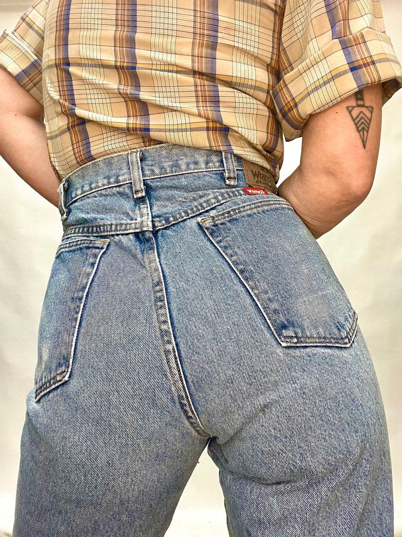 Op de afbeelding: Een lichtblauwe spijkerbroek met een hoge taille en een klassieke vijf-zak stijl. De spijkerbroek heeft een vervaagde wassing en een licht verweerde look. De achterzakken hebben een rood leren patch met het woord "Wrangler" erop gedrukt.