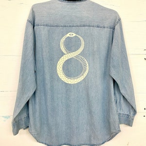 May include: A light blue denim shirt with a white snake design on the back. The snake is in the shape of the number eight and has an eye in the center of its head.