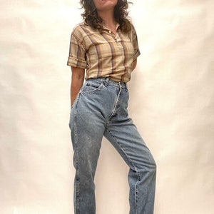 Op de afbeelding: Een vrouw draagt een lichtbruin en blauw geruit overhemd met korte mouwen en een lichtblauwe jeans met hoge taille.